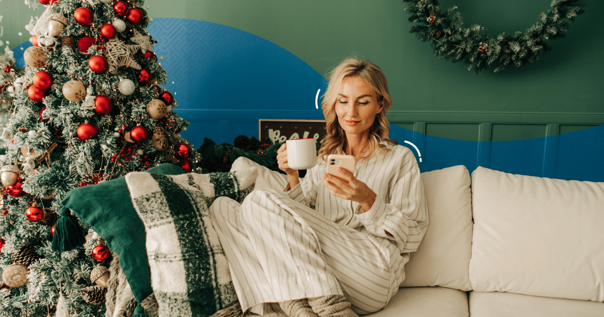 Woman sitting on couch drinking hot chocolate while texting on phone - How to Create Effective Holiday Text Marketing Campaigns